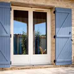 Volets battants rouges sur une maison en pierre traditionnelle, grande fenêtre bois et jardin fleuri avec rosiers et lavande. 2