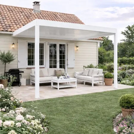 Pergola aluminium blanche adossée protégeant un salon de jardin sur terrasse avec maison et jardin fleuri