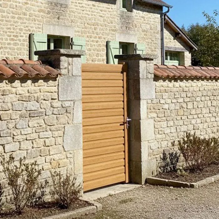 Portillon aluminium BAGO chêne doré devant un mur en pierre