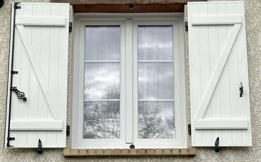 Volet battant aluminium blanc à deux vantaux ouverts, avec barres, écharpe, pentures noires et espagnolette, sur une fenêtre de maison à linteau bois