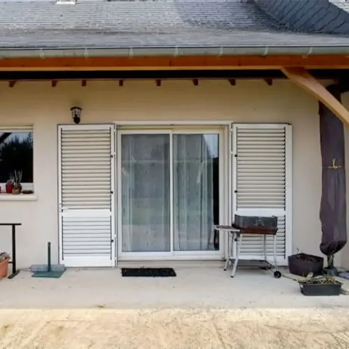 Volets battants persiennés blancs en aluminium de part et d'autre d'une baie coulissante, sur une maison avec terrasse
